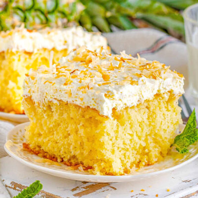 square piece of pina colada cake on a white plate frosted with white frosting and toasted coconut. a pineapple and another piece of cake can be seen in the background.