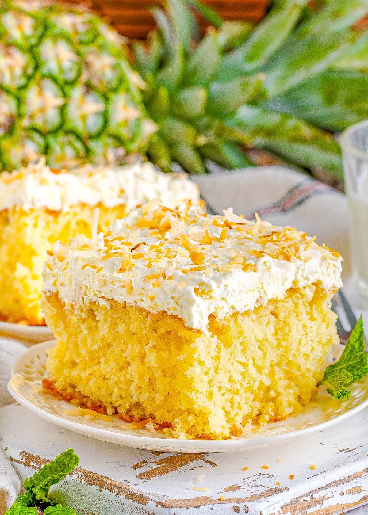 square piece of pina colada cake on a white plate frosted with white frosting and toasted coconut. a pineapple and another piece of cake can be seen in the background.
