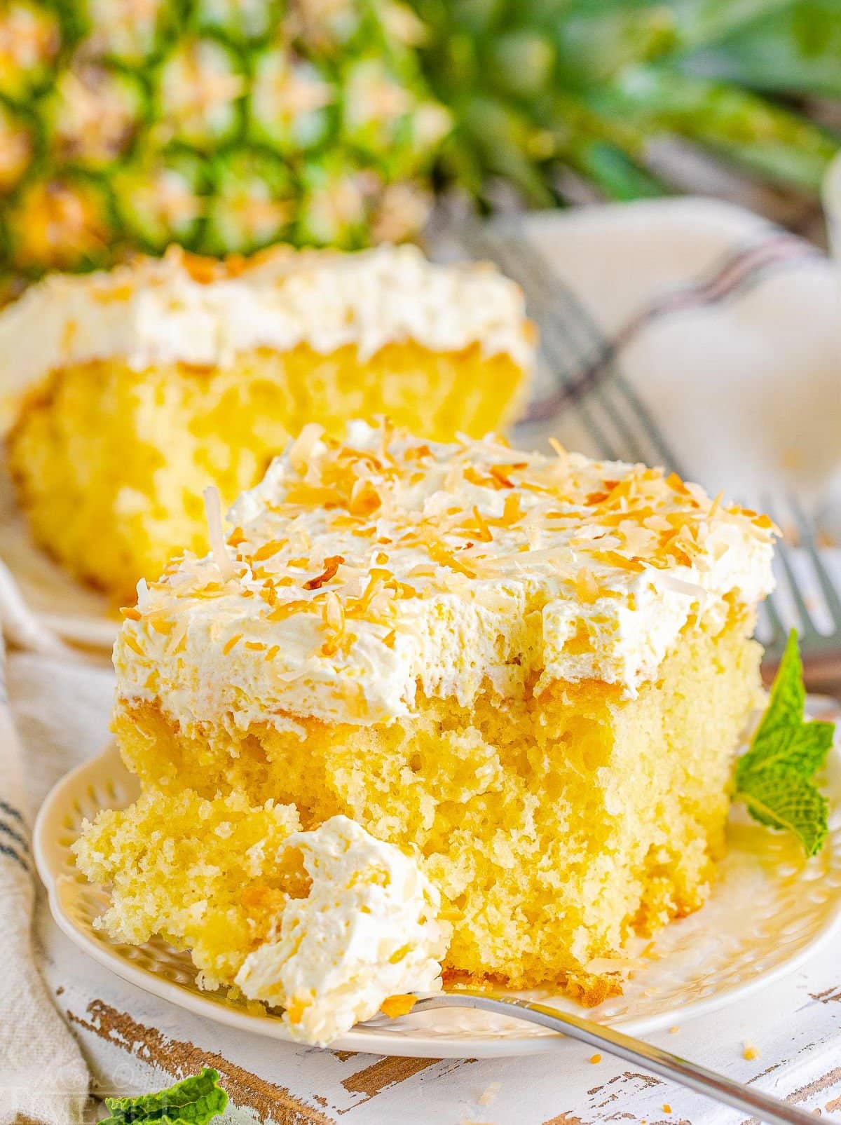 piece of pina colada poke cake on a white plate with a forkful of cake removed and sitting on edge of plate. another piece of cake and a whole pineapple can be seen in the background.