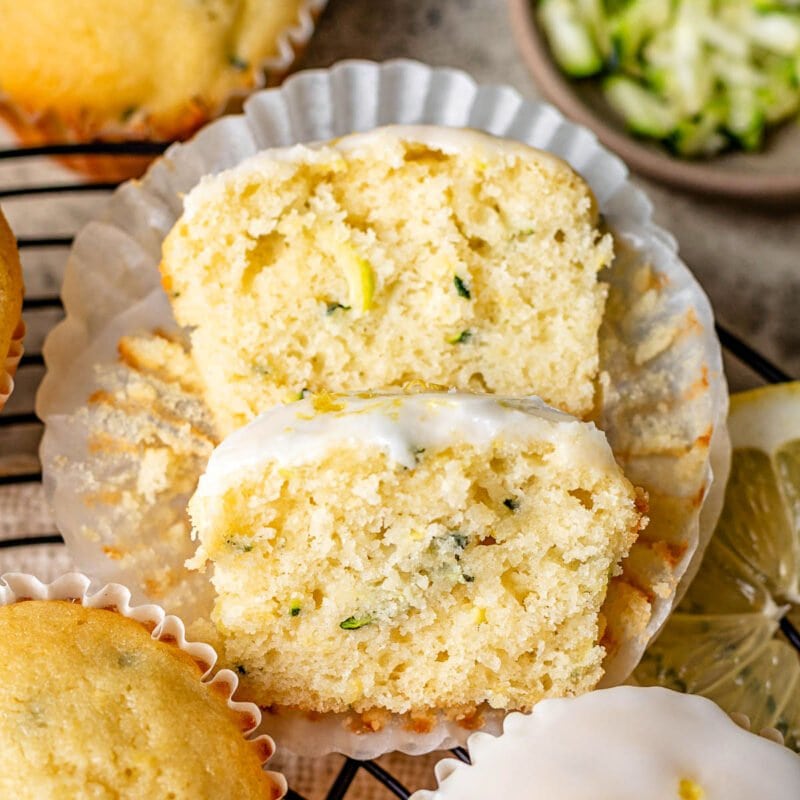 lemon zucchini muffin cut in half with one half resting on the other. three whole muffins and some shredded zucchini are near the cut muffin.