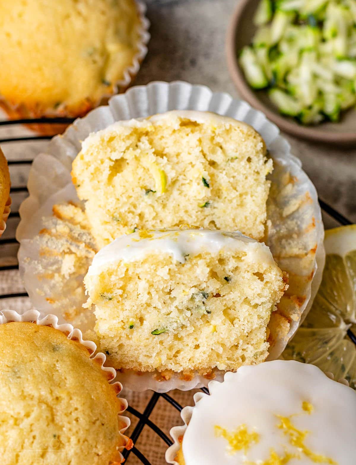 lemon zucchini muffin cut in half with one half resting on the other. three whole muffins and some shredded zucchini are near the cut muffin.