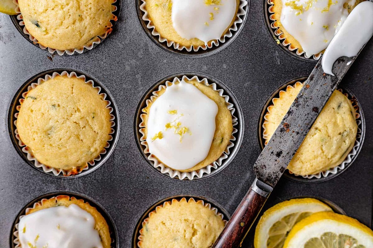 lemon zucchini muffins in muffin tin with half of them glazed and the glaze knife resting across the top of the pan.