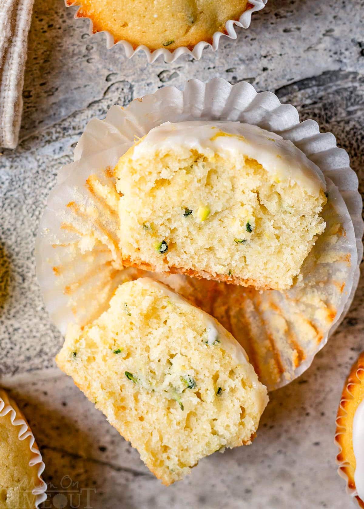 lemon zucchini muffin in white liner topped with lemon glaze cut in half and sitting on a gray backdrop.