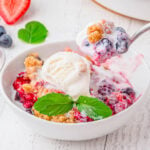 white bowl with berry cobbler in it topped with vanilla ice cream. spoonful of cobbler being scooped out.