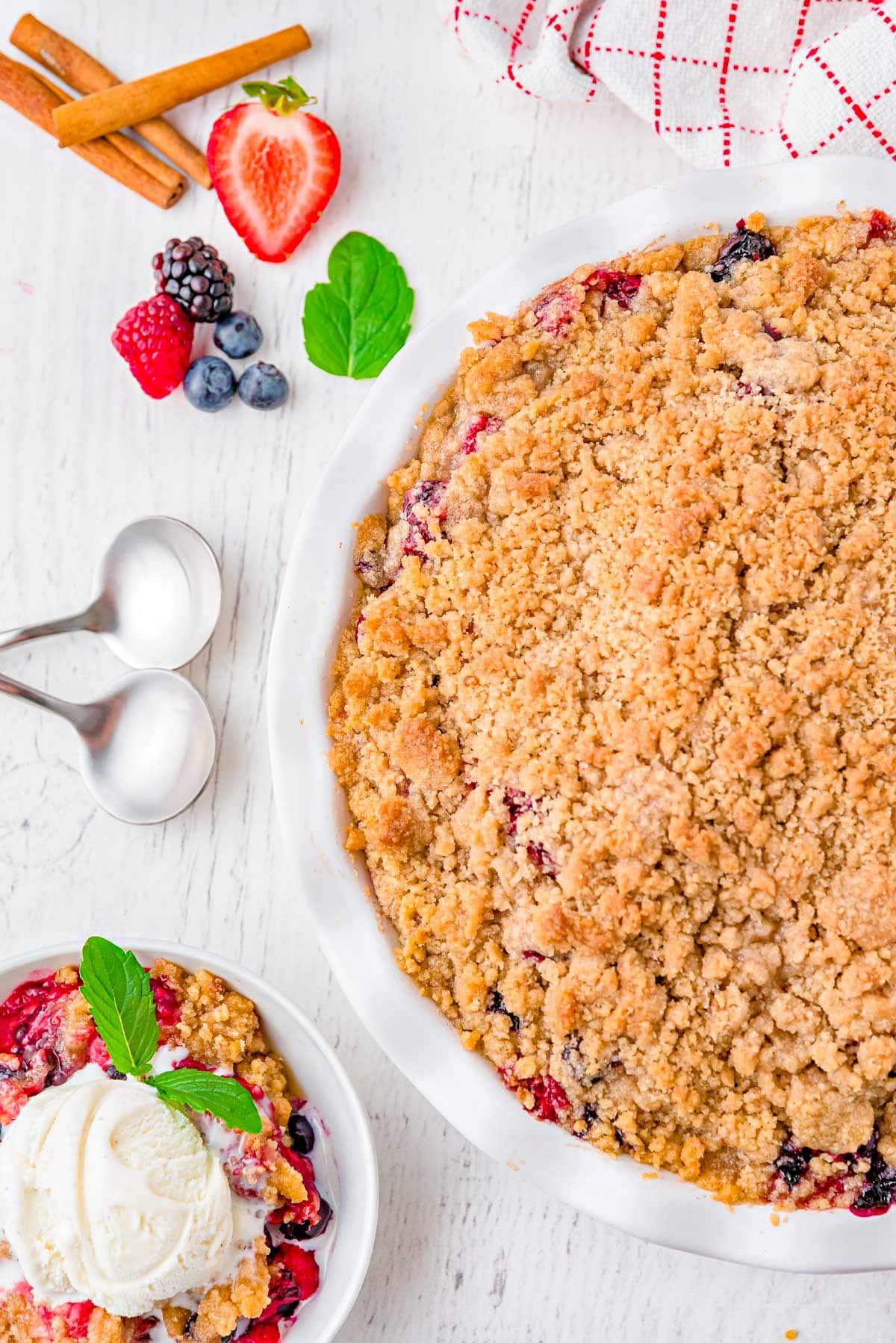 top down look at berry crumble in white pie plate with a serving removed and in a white bowl next to it. the crumble is topped with vanilla ice cream.