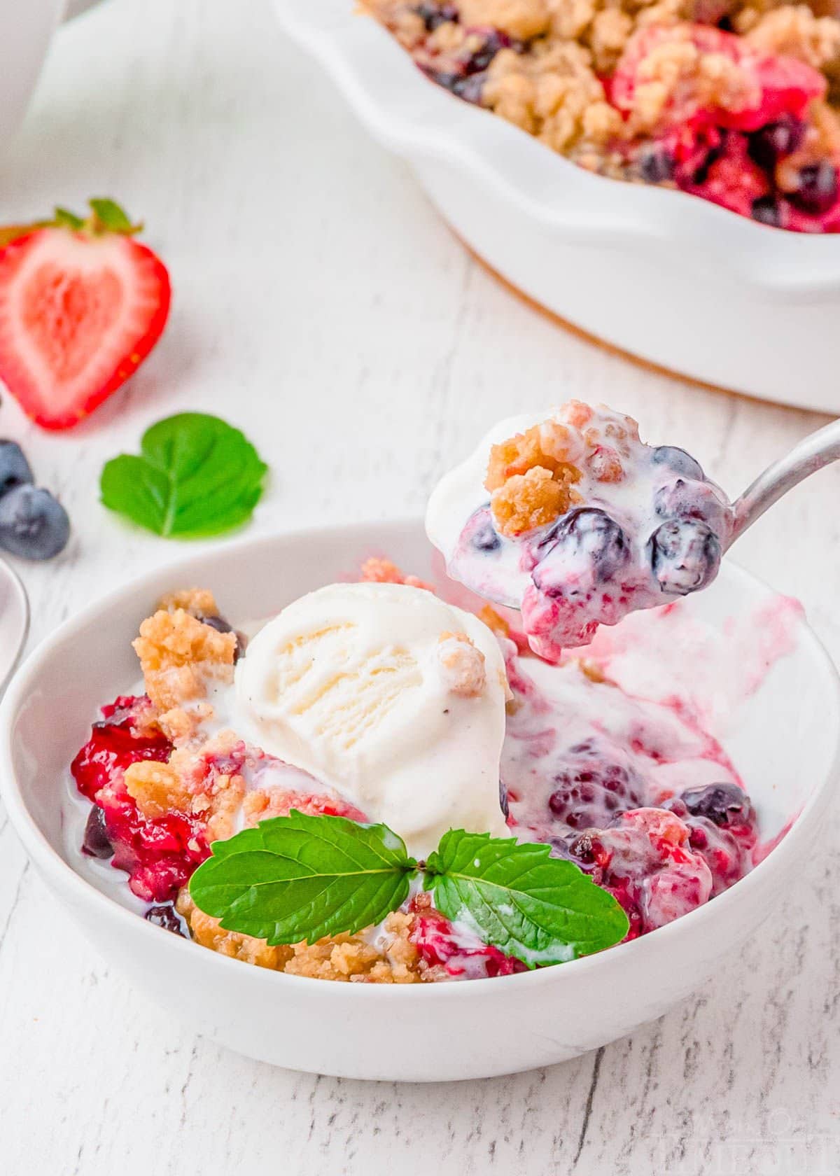 white bowl with berry cobbler in it topped with vanilla ice cream. spoonful of cobbler being scooped out.