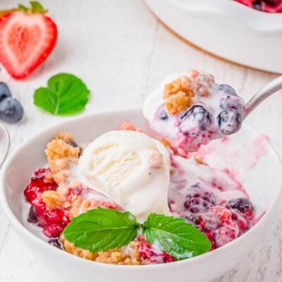 white bowl with berry cobbler in it topped with vanilla ice cream. spoonful of cobbler being scooped out.