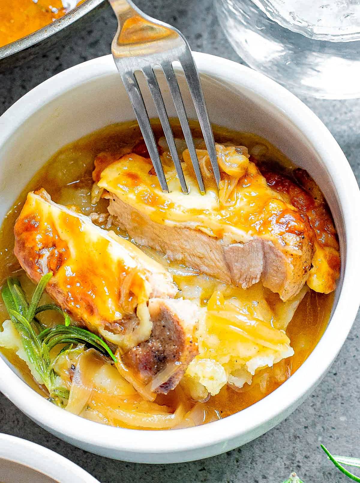 pork chop in small bowl on top of mashed potatoes cut in half so the juicy interior is showing. a fork is holding the two pieces apart.