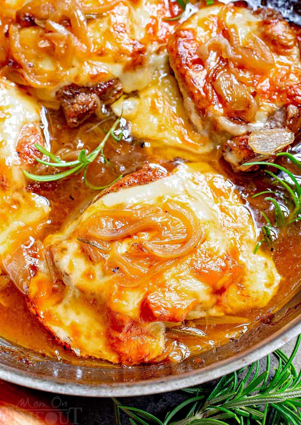 close up look at french onion pork chops in a stainless steel skillet topped with melted and browned cheese. fresh rosemary sprigs are scattered about.