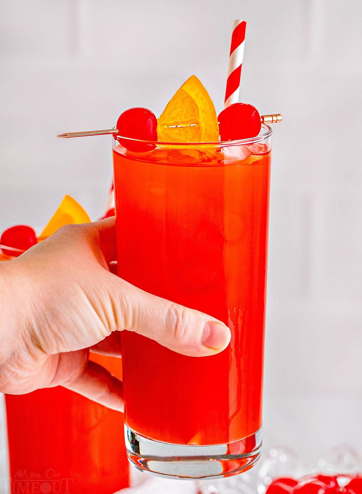 alabama slammer cocktail being held up in the air with a red and white striped straw with a maraschino cherry and orange garnish.