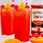 two tall glasses filled with alabama slammer recipe with red and white striped straws. bottle of southern comfort in the background and one orange has been cut in half.