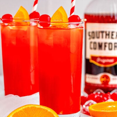 two tall glasses filled with alabama slammer recipe with red and white striped straws. bottle of southern comfort in the background and one orange has been cut in half.