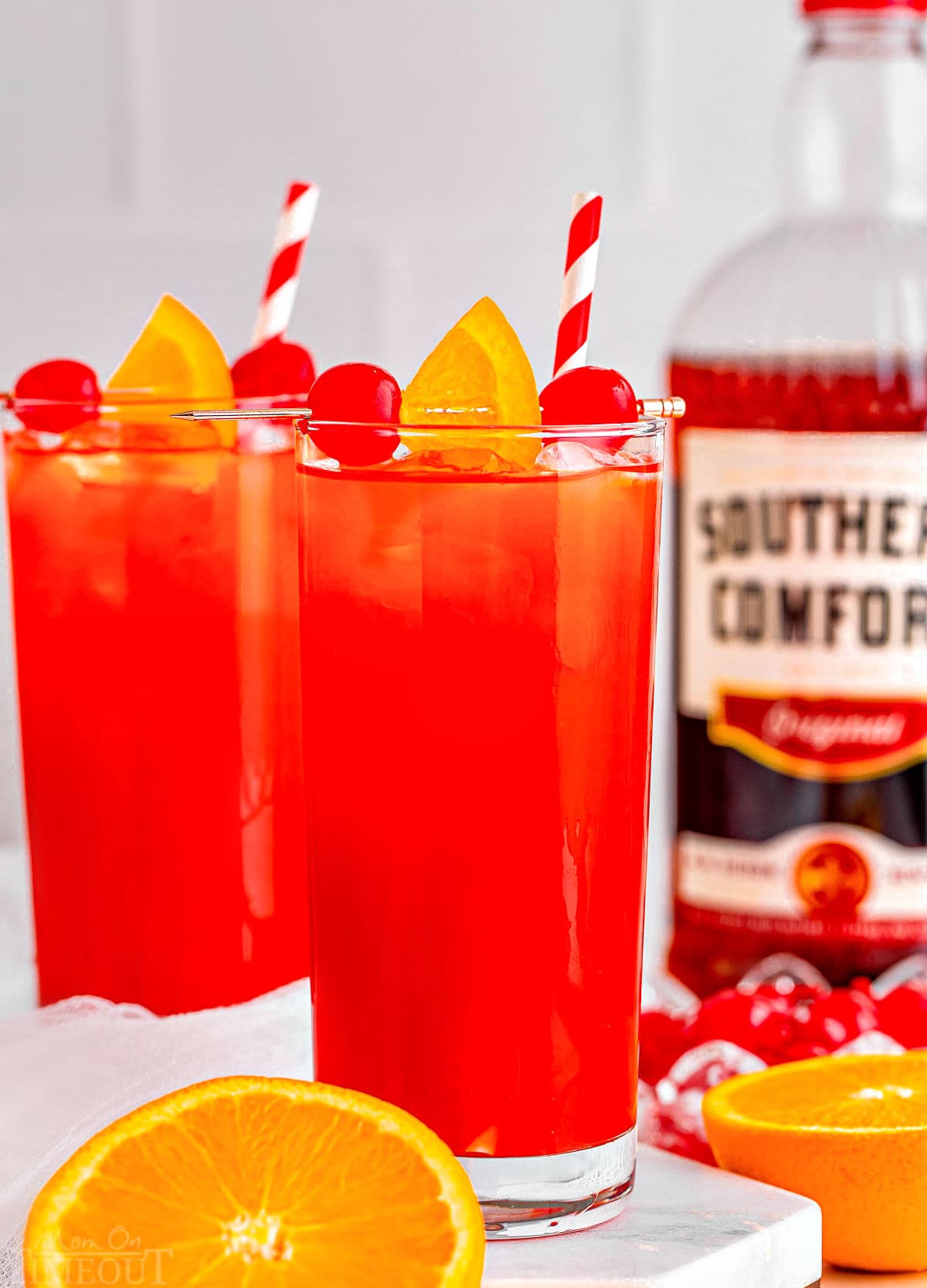 two tall glasses filled with alabama slammer recipe with red and white striped straws. bottle of southern comfort in the background and one orange has been cut in half.