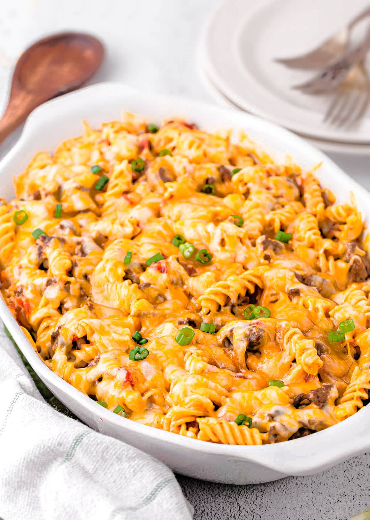 pasta casserole in white oval baking dish made with cheeseburger ingredients and topped with cheese. ready to be served onto small stack of plates in the background.