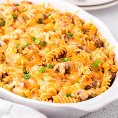 pasta casserole in white oval baking dish made with cheeseburger ingredients and topped with cheese. ready to be served onto small stack of plates in the background.