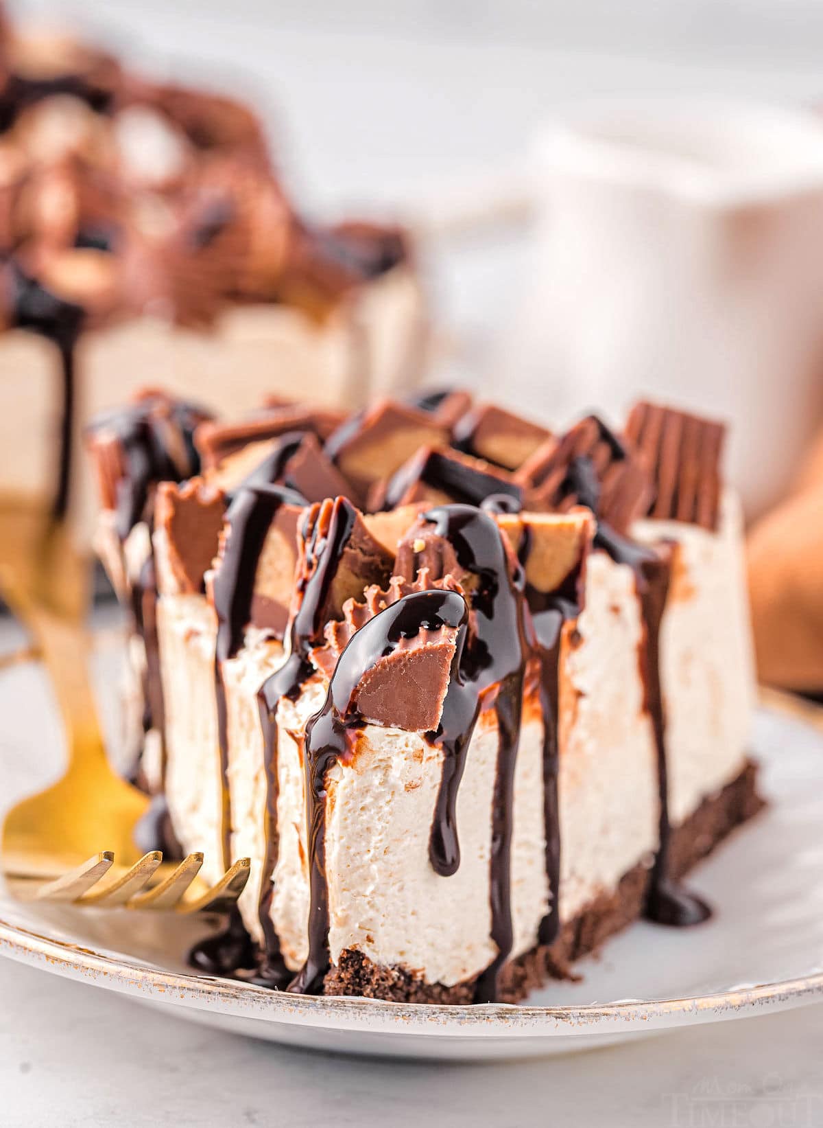 slice of no bake peanut butter cheesecake topped with reese's candies and hot fudge sitting on small gold rimmed white plate with a gold fork next to it.