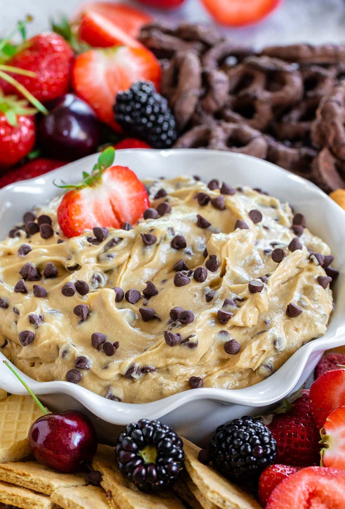 bowl with fluted edges holds chocolate chip cookie dough dip with half a strawberry dipped into the dip. Fresh fruit and cookies surround the bowl.