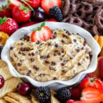 bowl with fluted edges holds chocolate chip cookie dough dip with half a strawberry dipped into the dip. Fresh fruit and cookies surround the bowl.