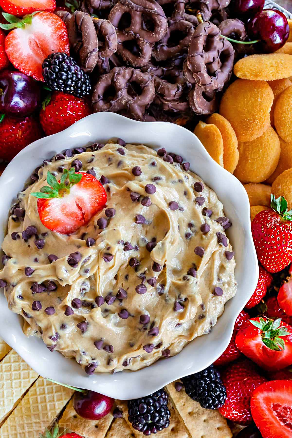 top down look at cookie dough dip in white bowl surrounded by strawberries, chocolate dipped pretzels, nilla wafers and other fruit and cookies.