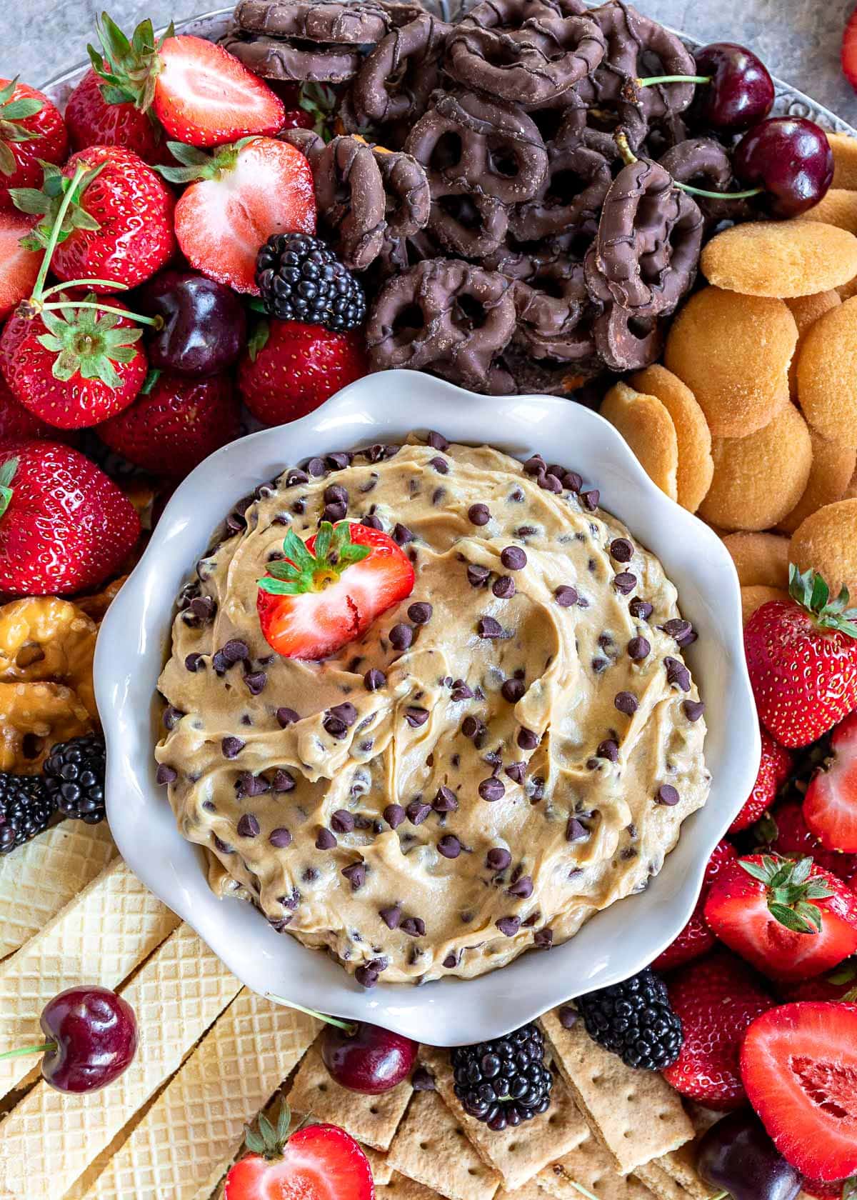 top down look at cookie dough dip in white bowl surrounded by strawberries, chocolate dipped pretzels, nilla wafers and other fruit and cookies. 