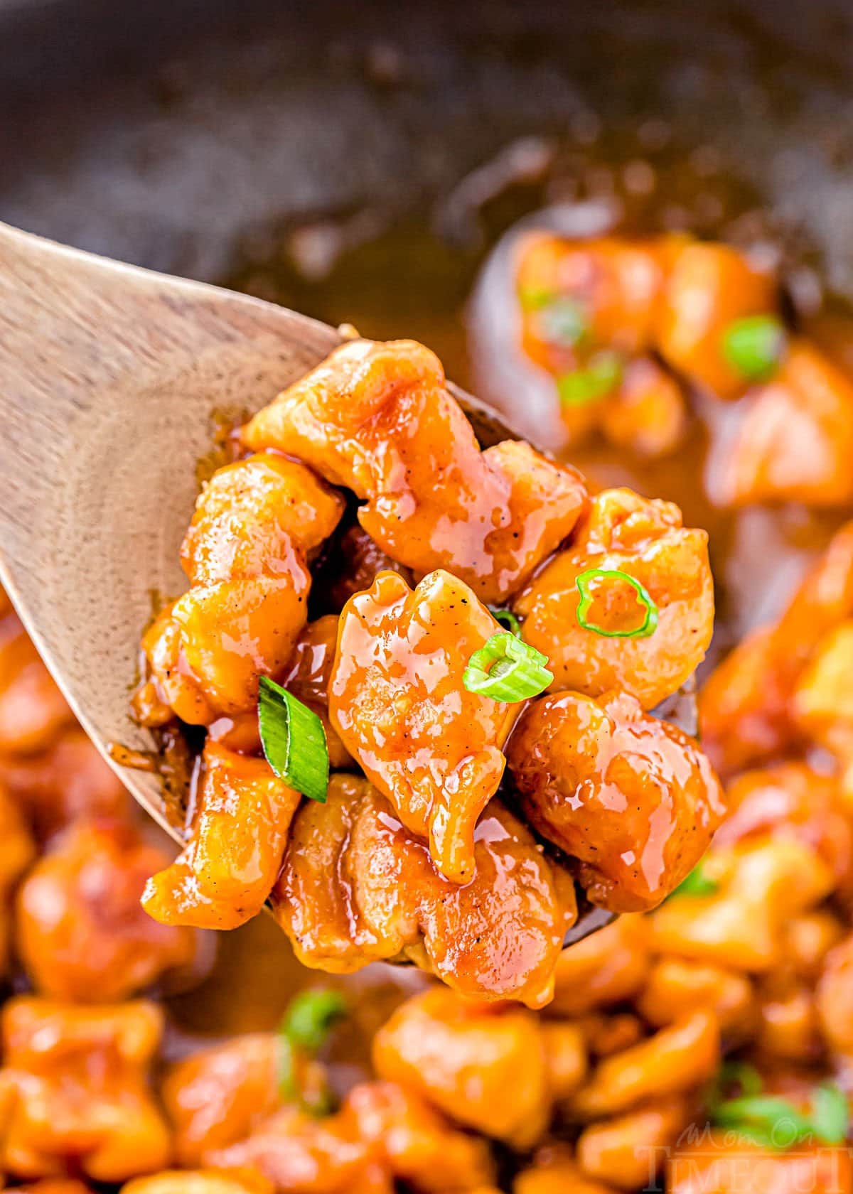 wooden spoon held above skillet with honey garlic chicken. garnished with sliced green onions.