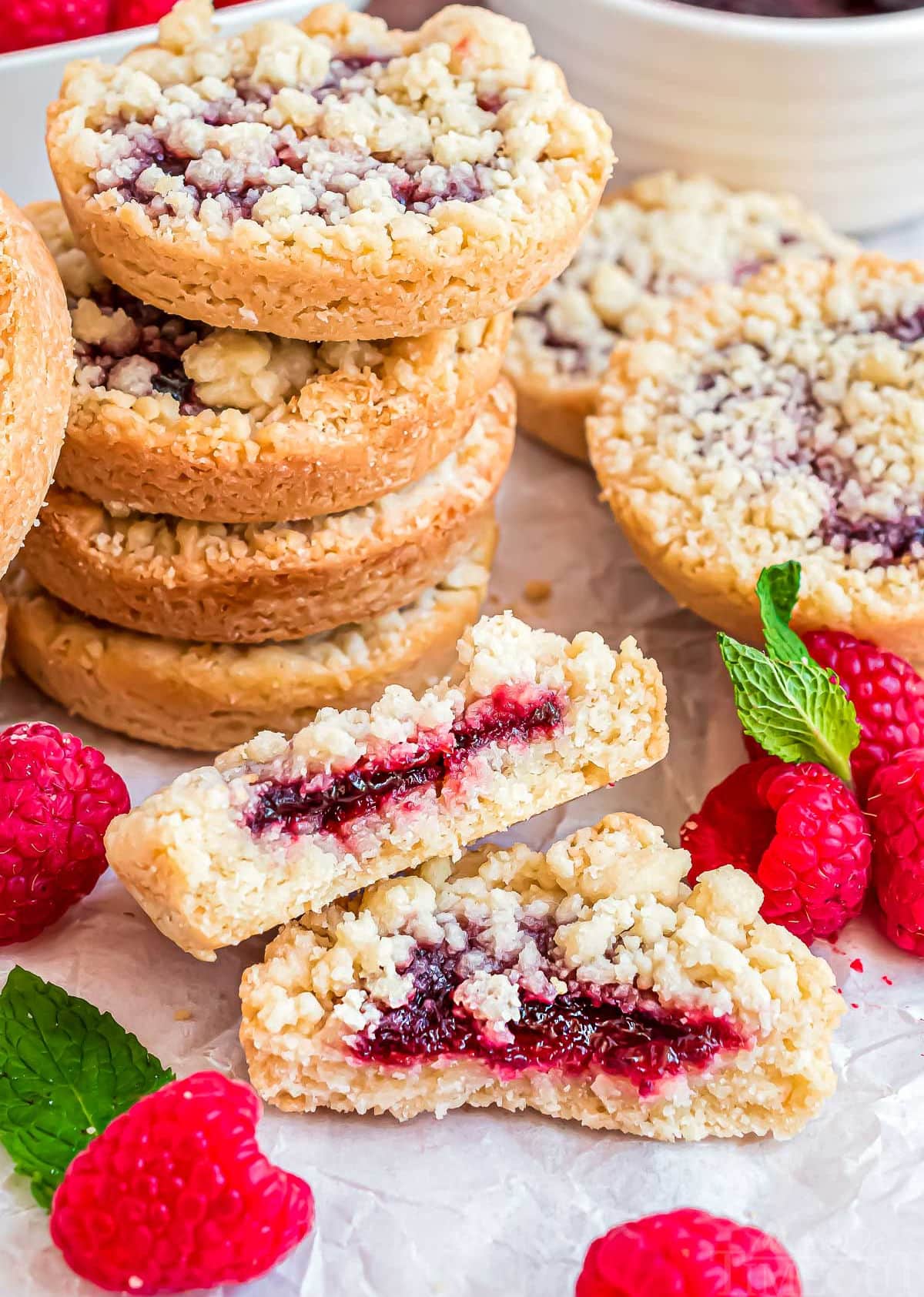 stack of mini raspberry crumble cookies with one cookie broken in half laying in front next to fresh raspberries.,