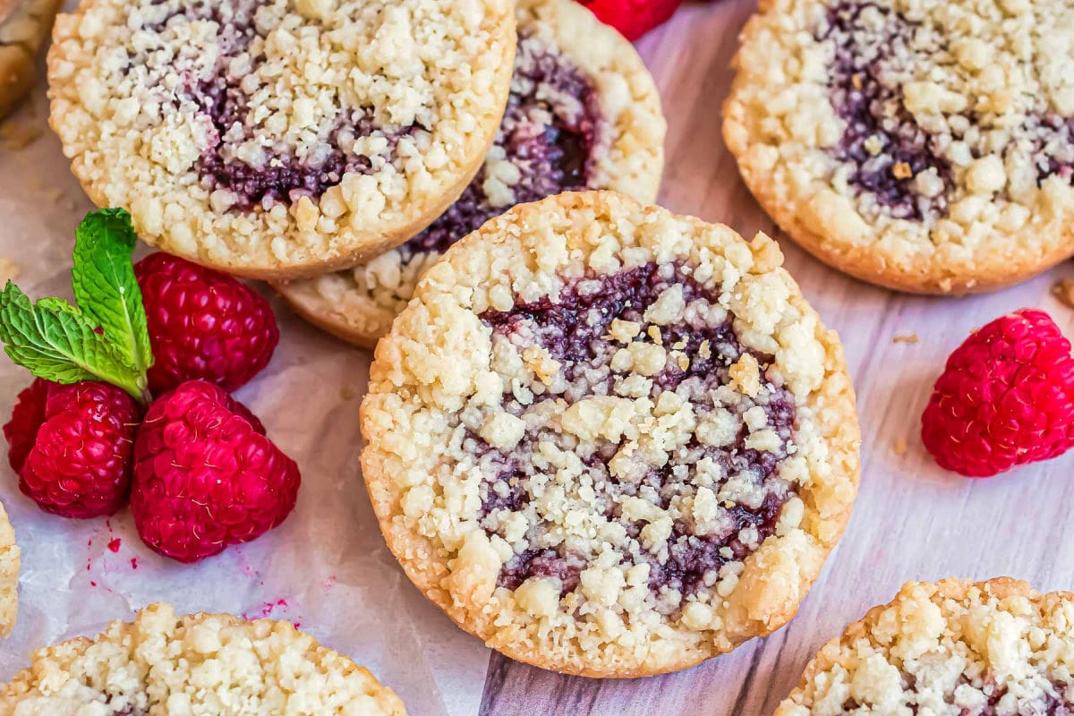 wide shot of top down look at raspberry crumble cookies laid out on white parchment with fresh mint and raspberries scattered about.