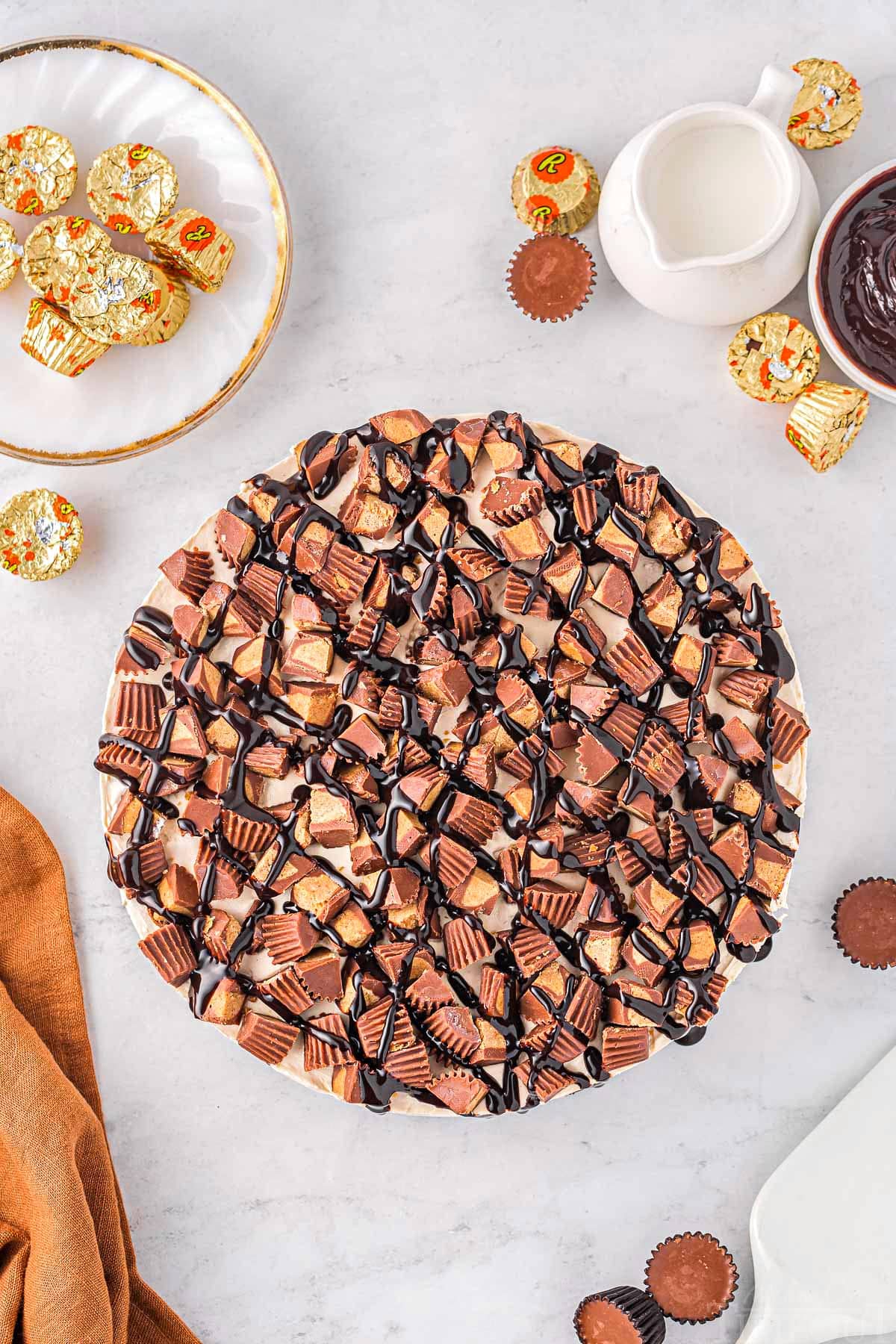 top down look at whole peanut butter cheesecake ready to be sliced and enjoyed. topped with reeses and hot fudge.