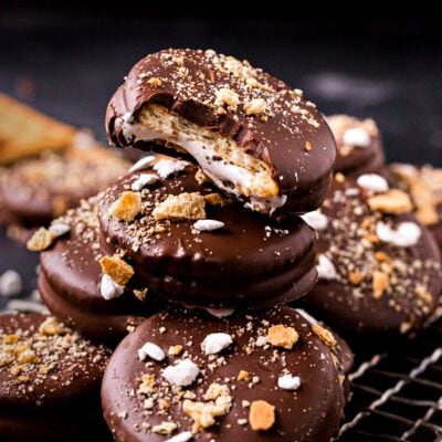 no bake ritz cracker sandwich cookies with marshmallow filling and dipped in chocolate stacked on small wire rack. tops of cookies have crushed graham crackers and miniature marshmallows.