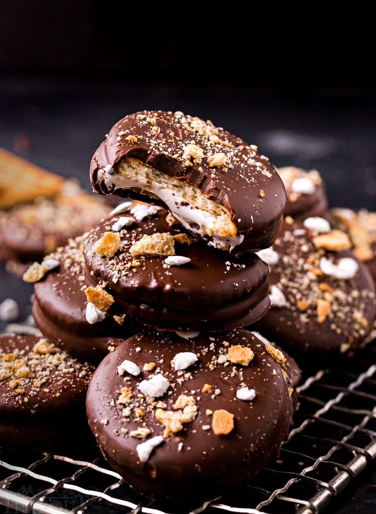 no bake ritz cracker sandwich cookies with marshmallow filling and dipped in chocolate stacked on small wire rack. tops of cookies have crushed graham crackers and miniature marshmallows.