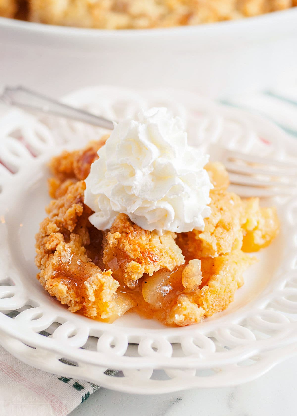 scoop of apple cobbler served on a white lace edged plate topped with fresh whipped cream. white casserole dish with remaining cobbler in the background.