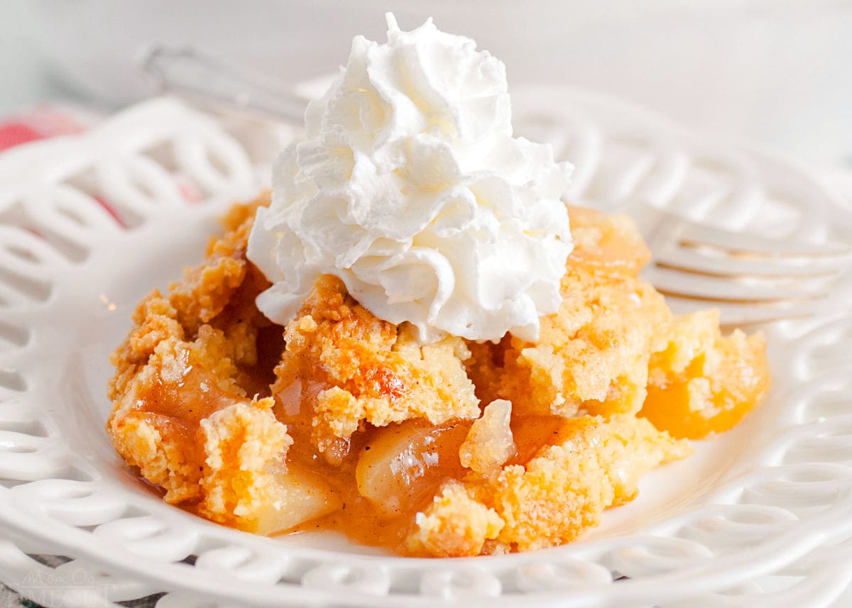 scoop of apple cobbler served on a white lace edged plate topped with fresh whipped cream. white casserole dish with remaining cobbler in the background.