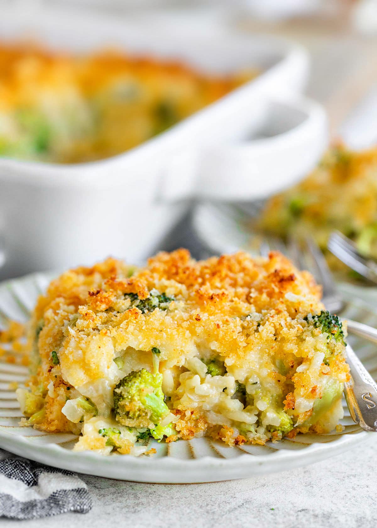 serving of broccoli rice casserole on a small decorative white plate with the casserole dish in the background.