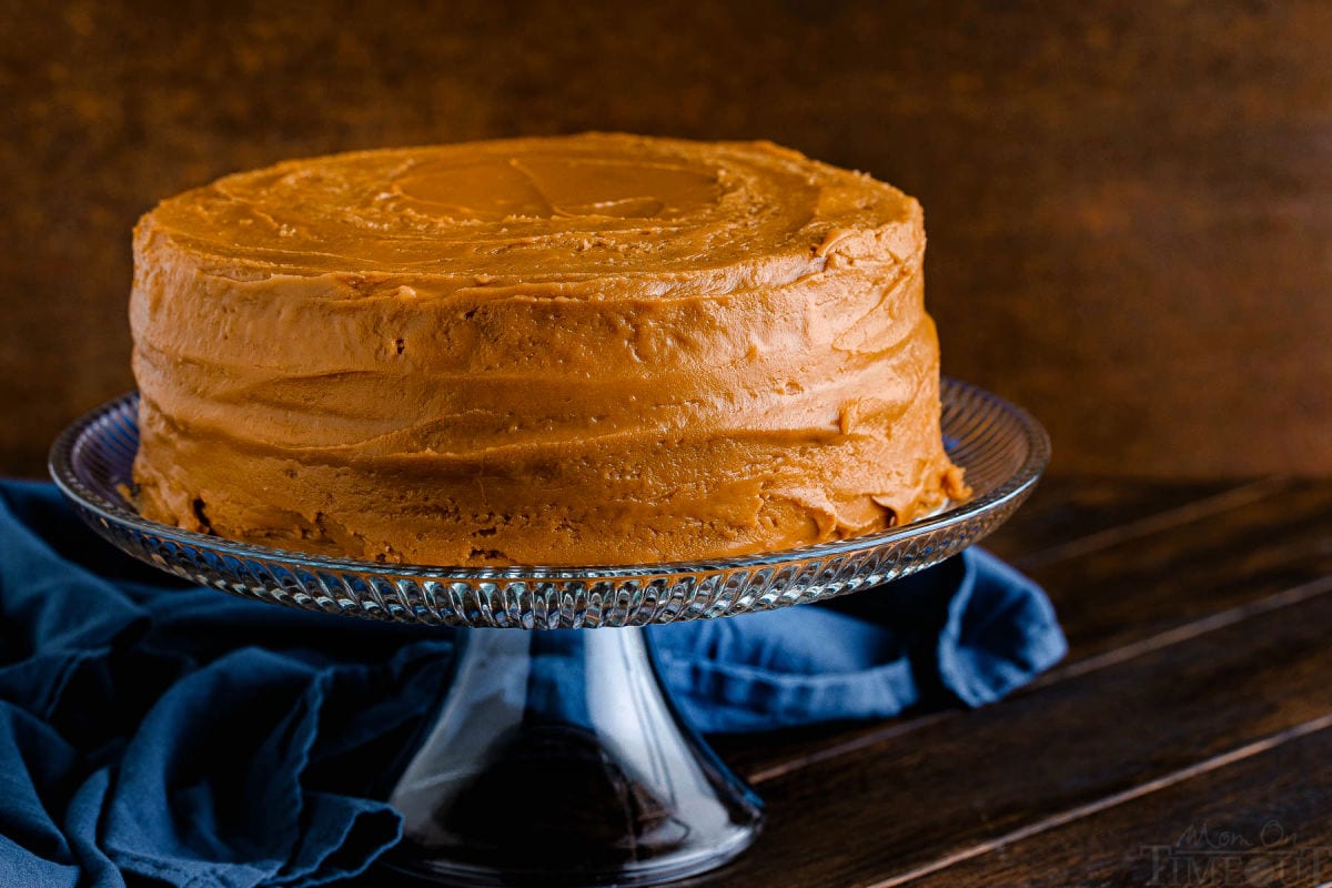 two layer cake with caramel frosting sitting on glass cake stand with a navy blue napkin in the background.