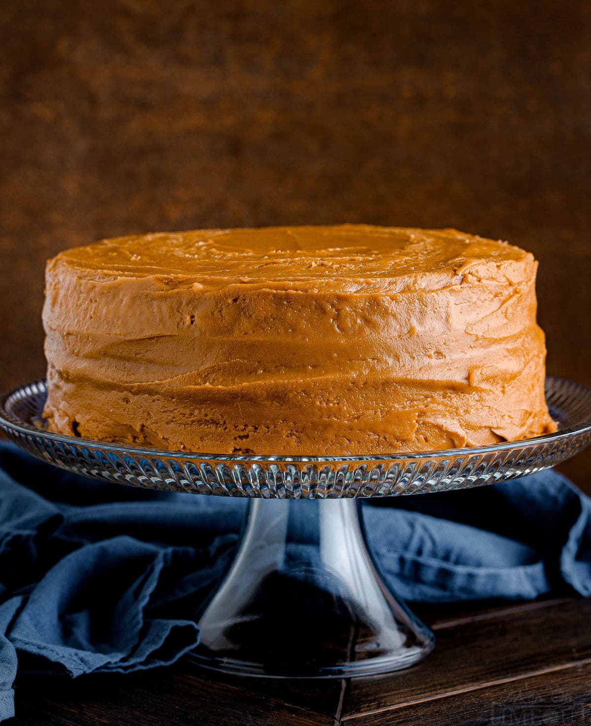 two layer cake with caramel frosting sitting on glass cake stand with a navy blue napkin in the backround.