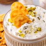 small clay bowl with dill pickle dip in it. three crackers are dipped into the dip with a sprinkle of chopped pickle scattered on top.
