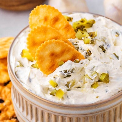 small clay bowl with dill pickle dip in it. three crackers are dipped into the dip with a sprinkle of chopped pickle scattered on top.