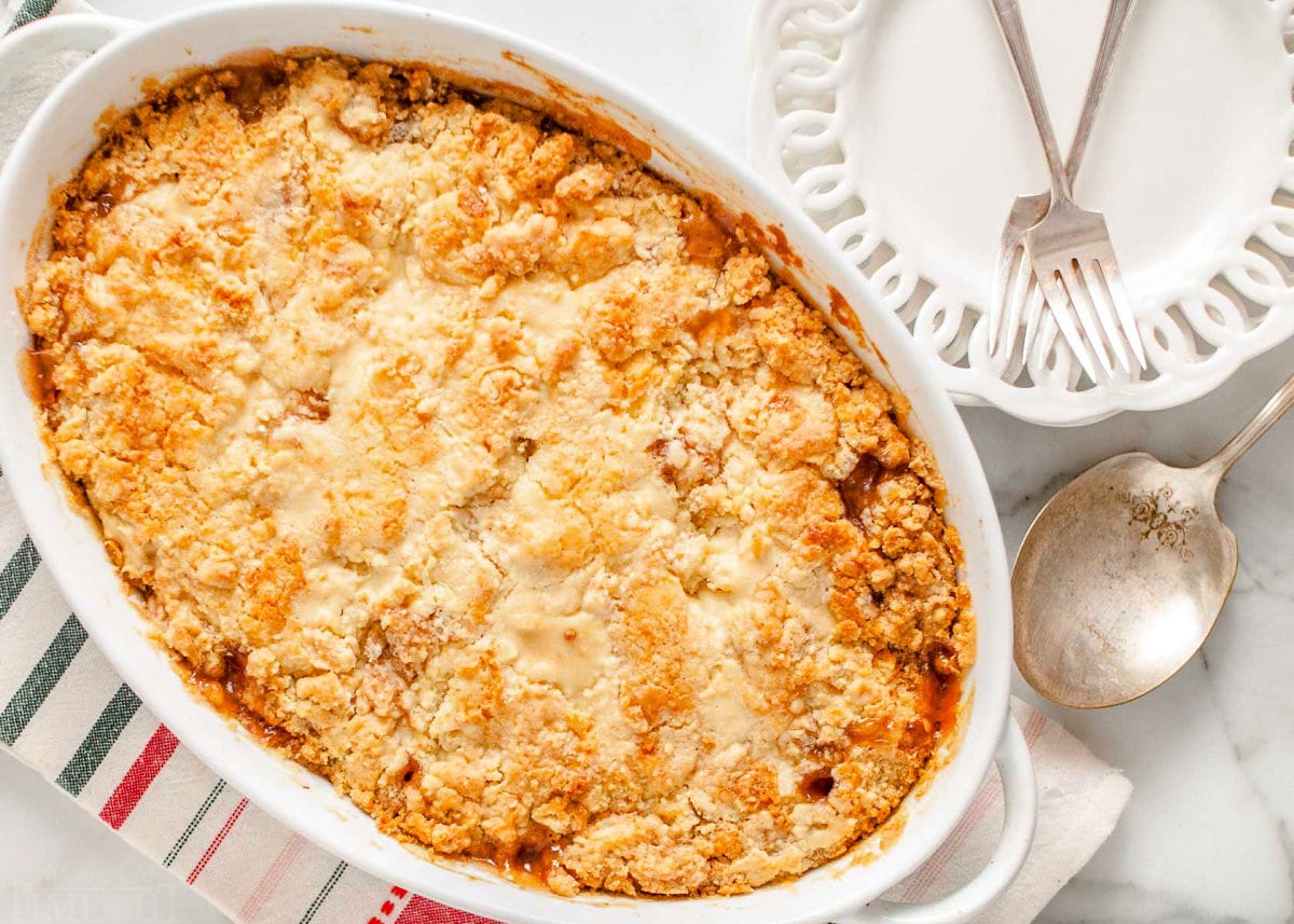 cooked apple cobbler in a white baking dish with two white plates and fork sitting off to the side.