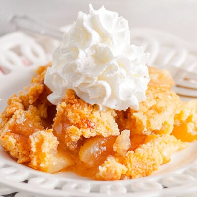 scoop of apple cobbler served on a white lace edged plate topped with fresh whipped cream. white casserole dish with remaining cobbler in the background.