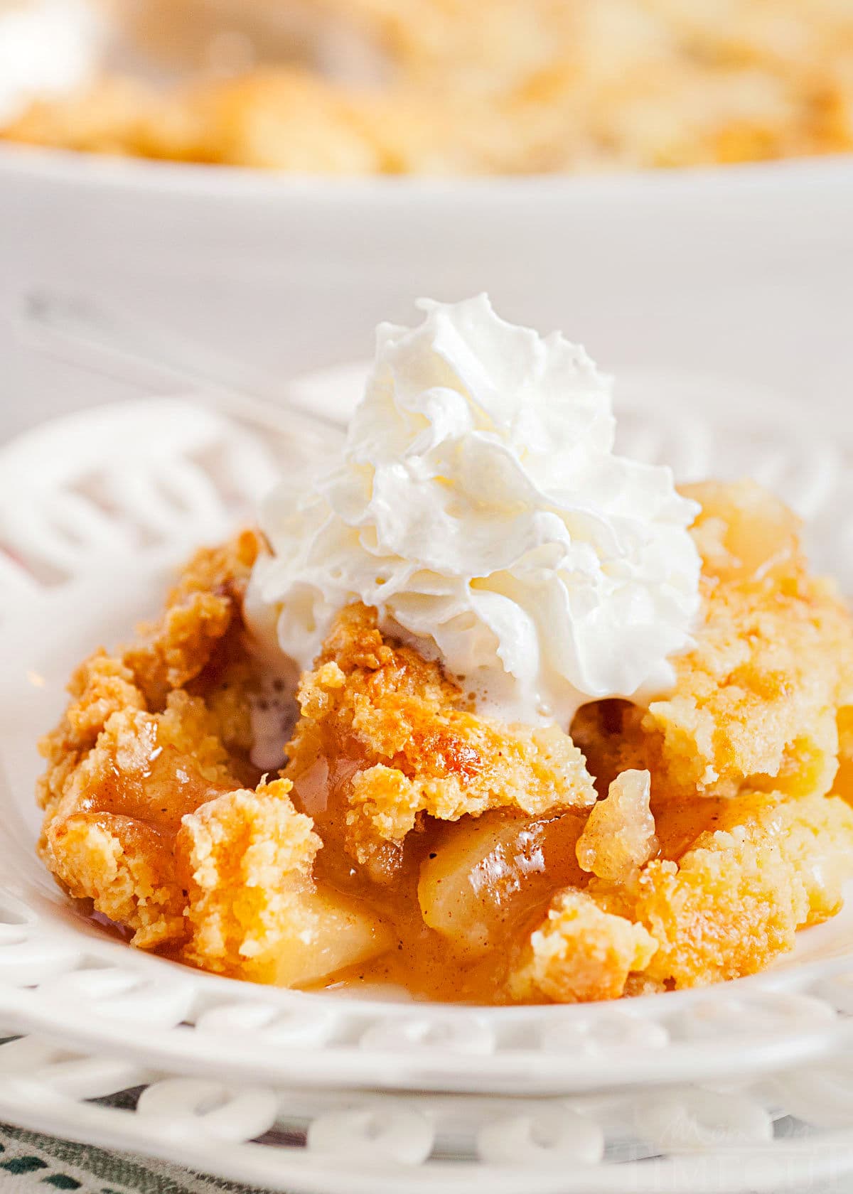 scoop of apple cobbler served on a white lace edged plate topped with fresh whipped cream. white casserole dish with remaining cobbler in the background.