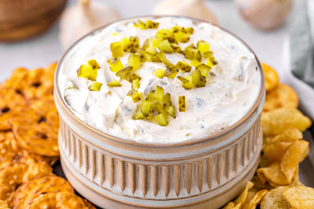 small decorative gray clay bowl with ridged detailing is filled with pickle dip and topped with more diced pickles. pretzels and chips surround the bowl.