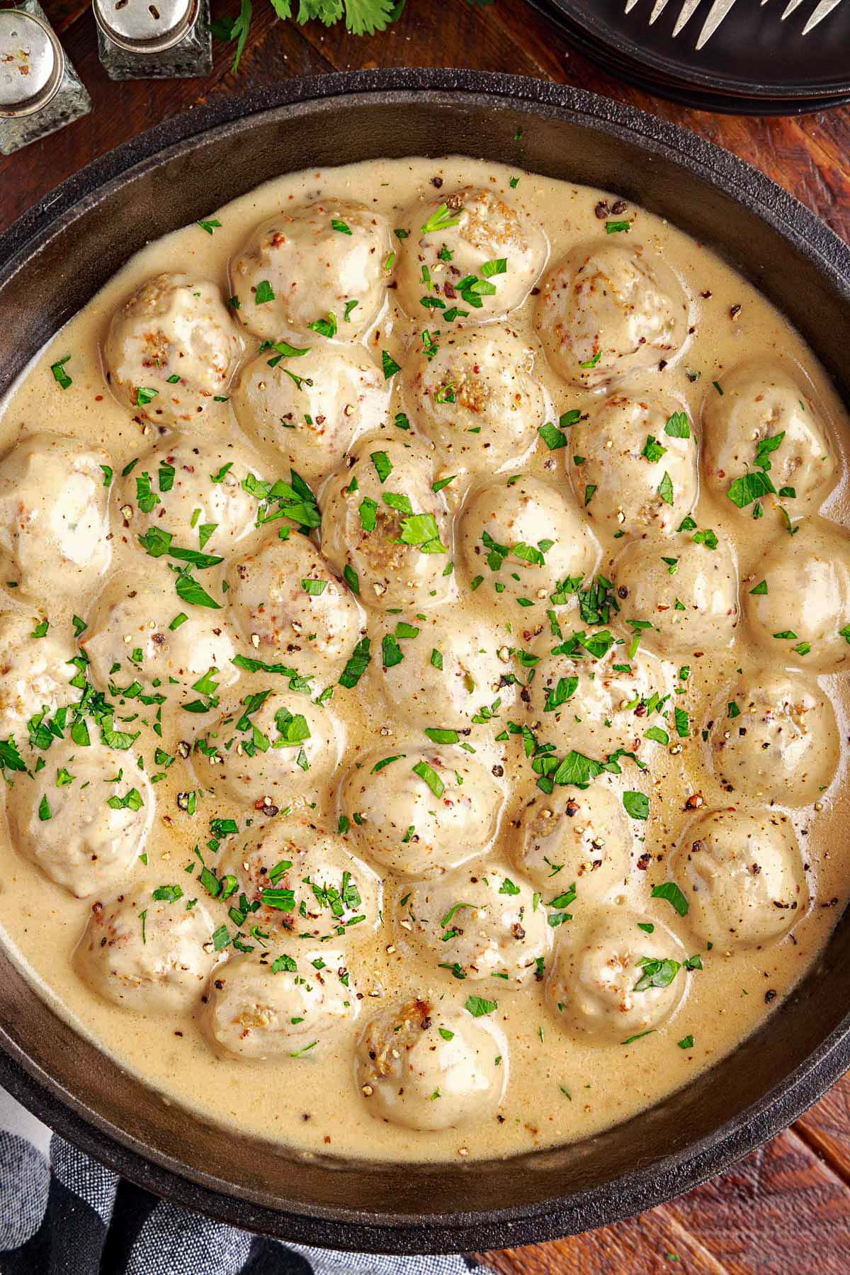 top down look at black cast iron skillet with swedish meatballs and gravy sitting on a dark wood surface with a black and white checked napkin nearby.