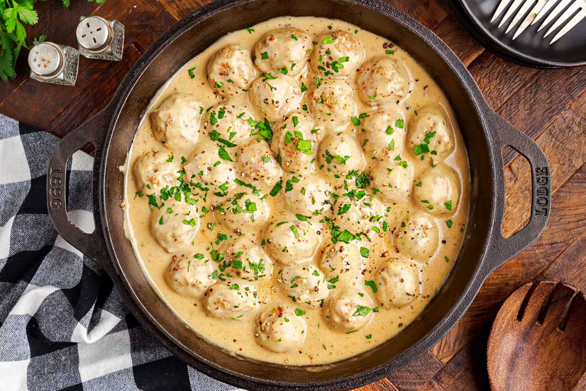 wide shot of a top down look at black cast iron skillet with swedish meatballs and gravy sitting on a dark wood surface with a black and white checked napkin nearby.