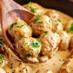 four meatballs on a wooden spoon being scooped out of skillet with the rest of the swedish meatballs and sauce.