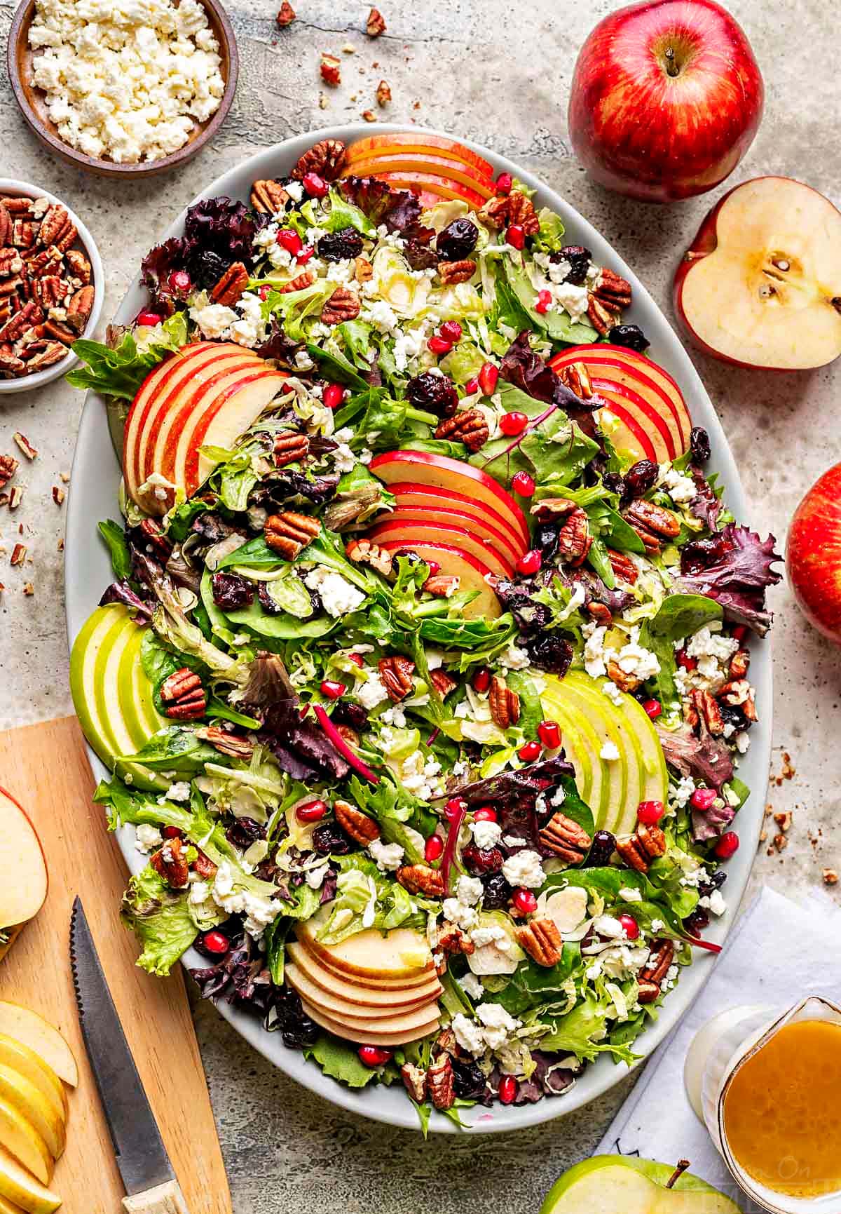 top down look at apple salad made with brussels sprout, apples, cranberries and topped with a vinaigrette. oval serving bowl is sitting on a gray surface with cut apples around it.