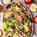 top down look at apple salad made with brussels sprout, apples, cranberries and topped with a vinaigrette. oval serving bowl is sitting on a gray surface with cut apples around it.