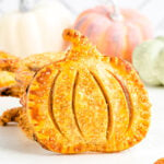 single pumpkin shaped pumpkin hand pie sitting upright in front of a stack of hand pies. an assortment of pumpkins can be seen in the background.