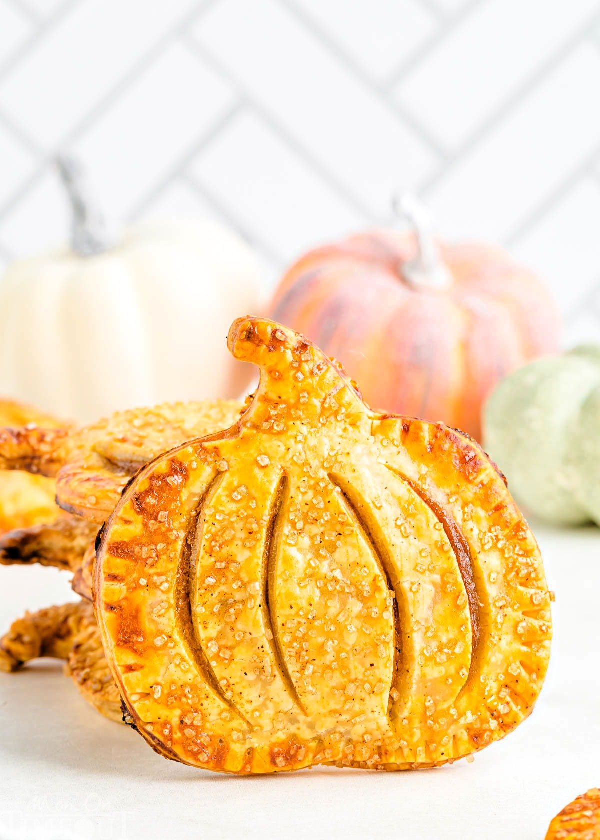 single pumpkin shaped pumpkin hand pie sitting upright in front of a stack of hand pies. an assortment of pumpkins can be seen in the background.