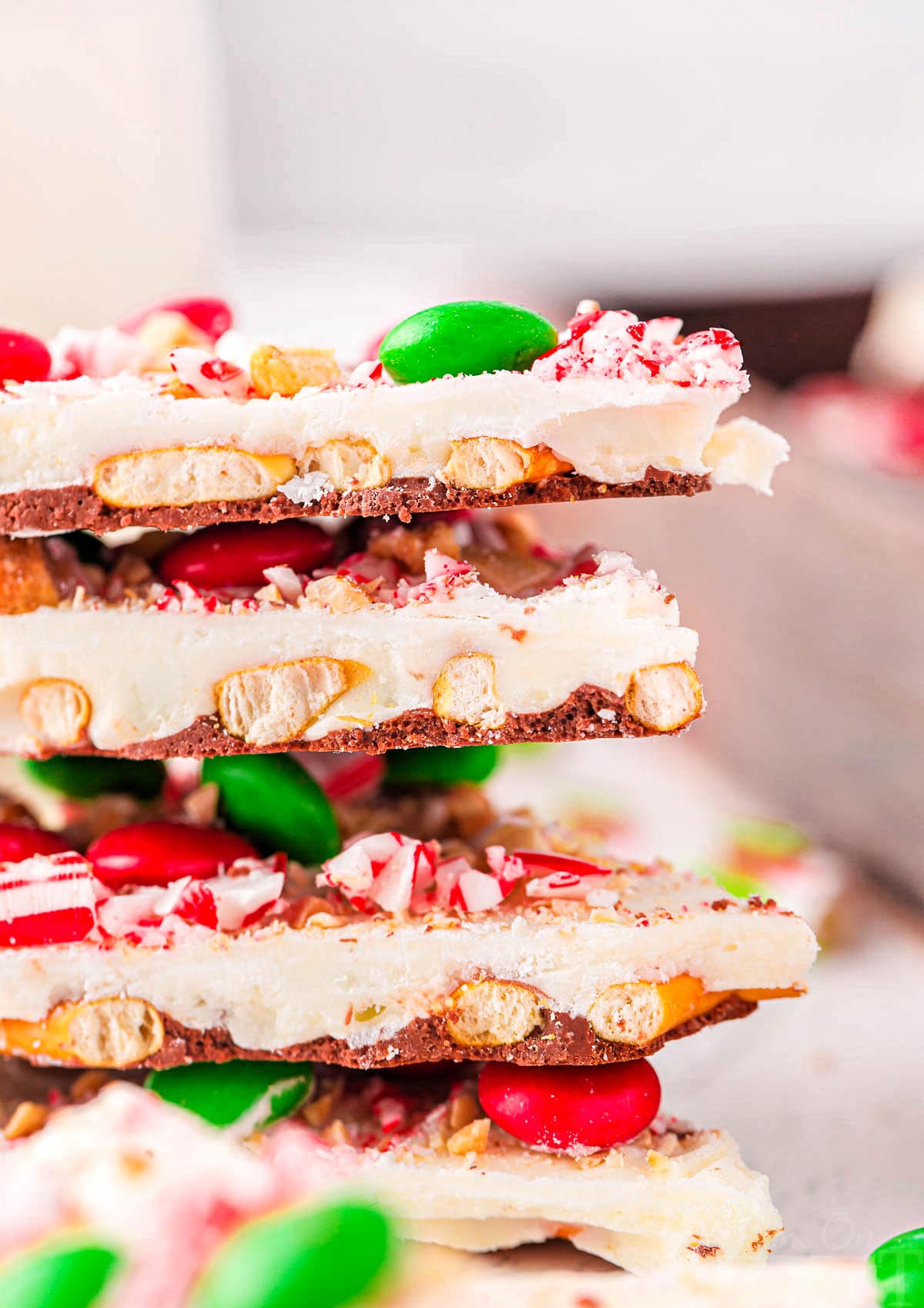 four pieces of christmas bark stacked on top of each other. the bark is made with pretzels, m and ms, and candy canes.