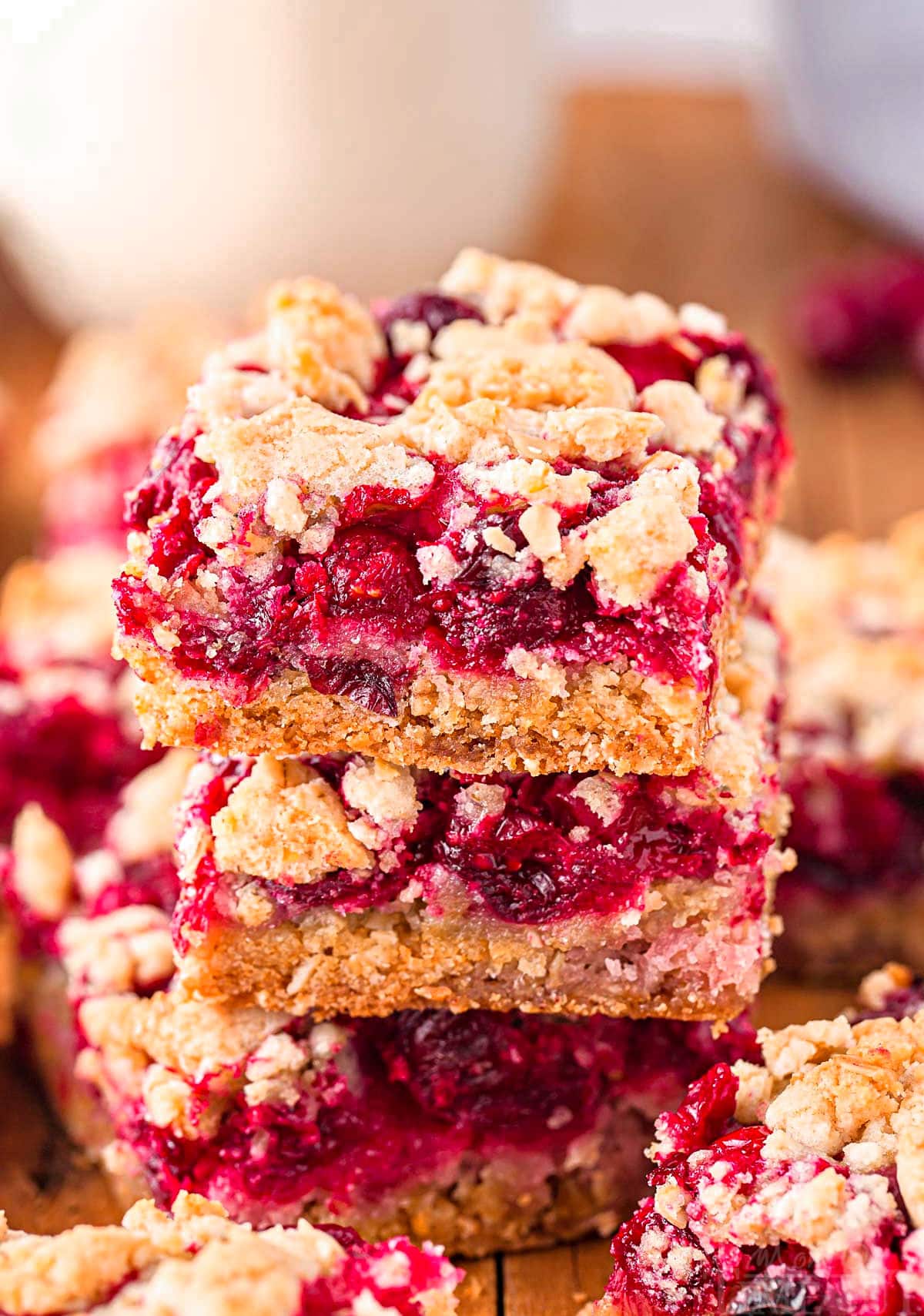 three cranberry crumb bars stacked on each other on a dark wood board with a glass of milk in the background.